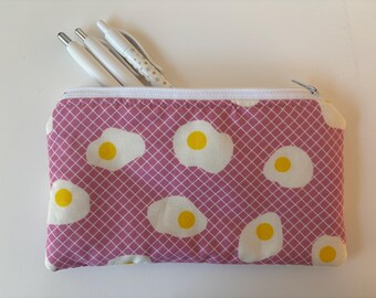 Peut inclure: Une trousse rectangulaire zippée avec un motif de grille rose et des motifs d'œufs au plat. Trois stylos blancs sont visibles à l'intérieur de la trousse ouverte. Les œufs ont des jaunes et des blancs d'œufs blancs.