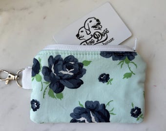 Peut inclure: Un petit porte-monnaie rectangulaire avec un motif floral. Le porte-monnaie est bleu clair avec des roses bleu marine et des feuilles vertes. Il a une fermeture éclair blanche, une dragonne blanche et un fermoir argenté. Une carte blanche avec un logo de chien est partiellement visible.