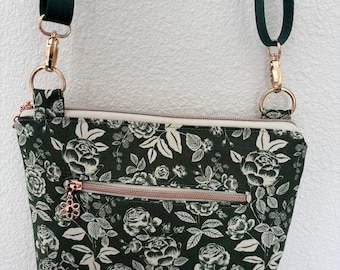 Peut inclure: Un sac bandoulière vert foncé avec un motif floral de roses et de feuilles blanches. Le sac a une fermeture éclair et des accessoires en or rose, et une bandoulière réglable vert foncé. Le sac est rectangulaire.