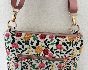 Peut inclure: Sac bandoulière à motifs floraux avec un fond blanc et un motif de roses roses, jaunes et rouges. Le sac a une sangle rose, une quincaillerie dorée et une tirette de fermeture éclair en forme de fleur. Il a deux compartiments zippés.