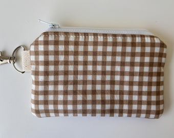 Peut inclure: Un petit porte-monnaie rectangulaire avec un motif vichy marron et blanc. Il est doté d'une fermeture éclair blanche et d'un fermoir en métal argenté fixé à une boucle en tissu beige. Le sac est conçu pour transporter de petits objets.