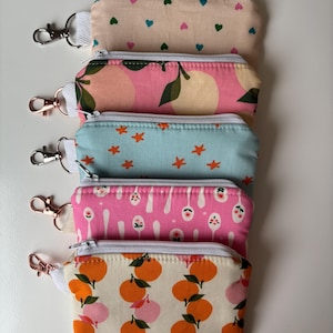 May include: Five rectangular fabric pouches with white zippers and metal lobster clasps. The top pouch is beige with small hearts, followed by a pink pouch with lemons, a light blue one with orange stars, a pink one with spoons, and a beige one with oranges.