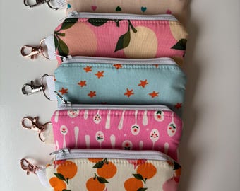 May include: Five rectangular fabric pouches with white zippers and metal lobster clasps. The top pouch is beige with small hearts, followed by a pink pouch with lemons, a light blue one with orange stars, a pink one with spoons, and a beige one with oranges.