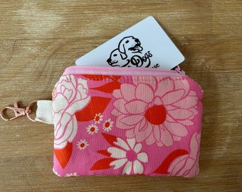 Peut inclure: Un petit portefeuille floral zippé avec un motif de fleurs roses et oranges. Le portefeuille a une sangle blanche avec un fermoir en or rose. Une carte blanche avec un logo de chien est partiellement visible.
