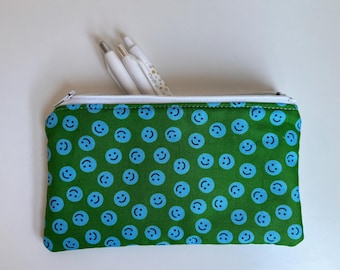 Peut inclure: Une trousse verte avec un motif répété de visages souriants bleus. La trousse a une fermeture éclair blanche et contient trois stylos blancs. La trousse est rectangulaire et en tissu.