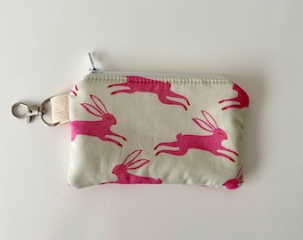 Peut inclure: Un petit porte-monnaie rectangulaire avec un fond crème et un motif répété de lapins roses vifs en mouvement. Le porte-monnaie a une fermeture éclair argentée, une boucle en tissu crème et un fermoir argenté.