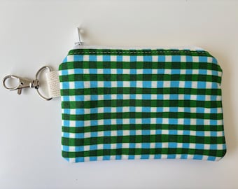 Peut inclure: Un petit porte-monnaie rectangulaire avec un motif à carreaux vert, bleu et blanc. Il possède une fermeture éclair blanche et un fermoir en métal argenté attaché à une boucle en tissu beige. Le porte-monnaie est probablement en tissu.