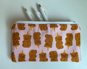 Peut inclure: Trousse rectangulaire avec un motif répétitif de glaces en forme d'ours bruns sur fond rose. La trousse a une fermeture éclair blanche et contient trois stylos blancs.