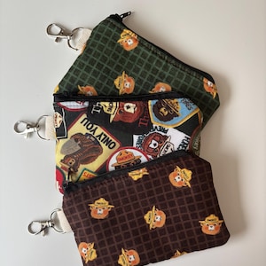 May include: Three small zippered pouches with attached keychains. The pouches feature different patterns, including a dark green grid with Smokey Bear, a black background with Smokey Bear patches, and a brown grid with Smokey Bear. The pouches have black zippers.