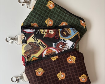 Peut inclure: Trois petites pochettes zippées avec porte-clés. Les pochettes présentent différents motifs, dont un quadrillage vert foncé avec Smokey Bear, un fond noir avec des écussons Smokey Bear et un quadrillage marron avec Smokey Bear. Elles ont des fermetures éclair noires.