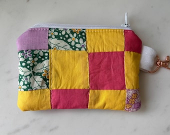 Peut inclure: Un porte-monnaie en patchwork avec une fermeture éclair blanche et un petit porte-clés. Le sac est fait de carrés de tissu jaunes, roses, verts et violets, avec des motifs floraux et géométriques. Il mesure environ 13 cm de large et 10 cm de haut.