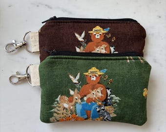 Peut inclure: Deux pochettes rectangulaires en tissu avec fermetures éclair noires et mousquetons en métal argenté. Une pochette est marron foncé, l'autre est vert forêt. Les deux présentent un imprimé d'un ours portant un chapeau marqué SMOKEY.
