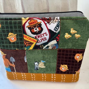 May include: A zippered pouch featuring a patchwork design with various Smokey Bear themed fabric squares in shades of green, brown, and mustard yellow. The fabric displays illustrations of Smokey Bear, the text Smokey's Helper, and forest animals.