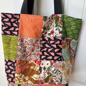 May include: A patchwork tote bag with a black strap. The bag features a variety of fabric squares in floral, bird, and geometric patterns. Colors include black, pink, orange, green, and brown. The bag is designed for carrying items.