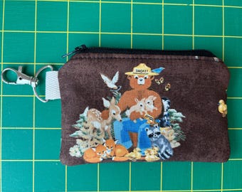 Peut inclure: Une pochette zippée en tissu marron avec une illustration imprimée de Smokey Bear portant un chapeau, entouré d'animaux de la forêt dont un cerf, un renard, un raton laveur et des oiseaux. Un mousqueton en métal argenté est fixé sur le côté.