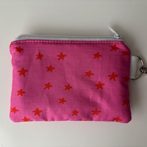 May include: A pink zippered pouch with a red star pattern. The pouch has a white zipper and a silver-colored clasp attached to a white fabric loop. The pouch is on a white surface.