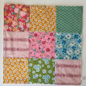 May include: A square patchwork quilt featuring nine distinct fabric panels with various floral and geometric patterns in shades of pink, mustard yellow, green, light blue, and white. The fabric squares are arranged in a three-by-three grid layout.