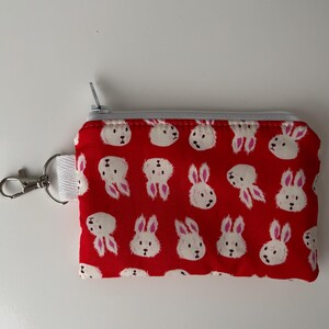 May include: Red fabric coin purse with a white bunny pattern. The small zippered pouch has a silver zipper and a white strap with a silver clasp. The bunny pattern features white bunnies with pink ears and noses.