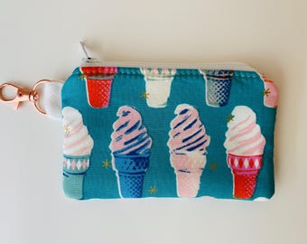 Peut inclure: Porte-monnaie turquoise avec un motif répété de cornets de glace roses, bleus et blancs. Le sac a une fermeture éclair et un fermoir or rose avec une sangle blanche. Les cornets de glace sont dans des cônes rouges, blancs et bleus.