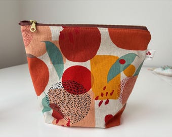 Peut inclure: Une trousse de maquillage en tissu avec un motif abstrait coloré. Le design présente de grandes formes rouges, oranges, jaunes et turquoise sur un fond crème. Elle a une fermeture éclair marron et une petite étiquette avec un logo.