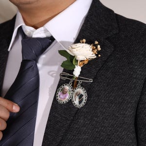 May include: A boutonniere featuring a white rose and small dried flowers, attached with a silver safety pin. Two oval photo pendants hang below, each with a decorative silver frame. The boutonniere is pinned to a dark gray suit jacket.
