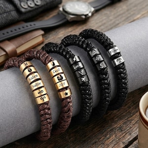 Könnte beinhalten: Vier geflochtene Armbänder mit personalisierten Metallperlen. Die Armbänder sind braun und schwarz, mit goldenen und schwarzen Perlen. Die Perlen sind mit Namen, Daten und Symbolen graviert. Eine Uhr und eine Lederbrieftasche sind im Hintergrund.