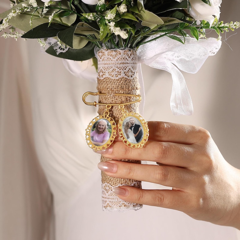 May include: A bridal bouquet with white flowers and greenery, adorned with two gold-colored memorial photo charms. The charms are attached to the bouquet with a safety pin. The bouquet's handle is wrapped in burlap and lace.