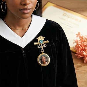 May include: A gold-colored graduation pin with a blue gemstone and the year 2026. The pin features a mortarboard and diploma design, with a circular locket holding a photograph. The pin is attached to a black graduation gown with a white collar.