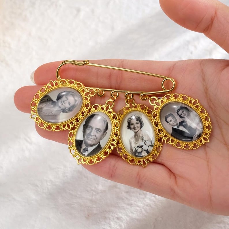 May include: A gold-colored brooch featuring four oval-shaped frames with black and white portraits. The frames are connected to a safety pin-style clasp. The portraits appear to be of people.