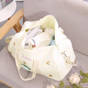 K&ouml;nnte beinhalten: Eine hellgelbe, gesteppte Tragetasche mit gestickten Teddyb&auml;ren und dem Namen "Leela" in Schreibschrift. Die Tasche ist offen und zeigt B&uuml;cher, eine Bommelm&uuml;tze und andere Gegenst&auml;nde. Sie hat einen Schultergurt und Griffe.