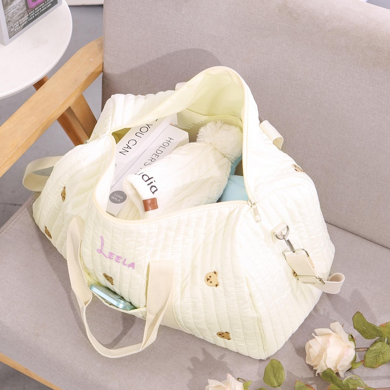 K&ouml;nnte beinhalten: Eine hellgelbe, gesteppte Tragetasche mit gestickten Teddyb&auml;ren und dem Namen "Leela" in Schreibschrift. Die Tasche ist offen und zeigt B&uuml;cher, eine Bommelm&uuml;tze und andere Gegenst&auml;nde. Sie hat einen Schultergurt und Griffe.