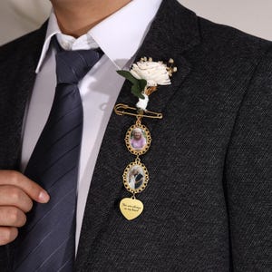 May include: A gold-colored memorial brooch with two oval photo frames and a heart-shaped charm. The heart charm has the inscription "You are always in my heart." A white flower and greenery are attached to the brooch.