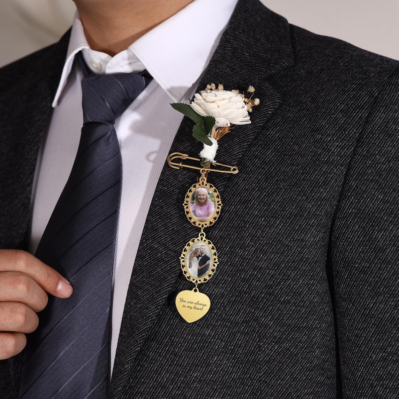 May include: A gold-colored memorial brooch with two oval photo frames and a heart-shaped charm. The heart charm has the inscription "You are always in my heart." A white flower and greenery are attached to the brooch.
