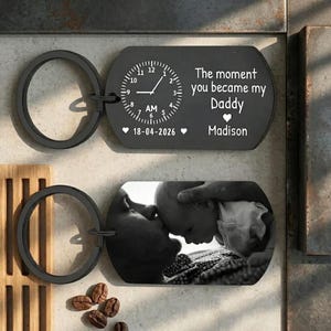 Op de afbeelding: Twee zwarte rechthoekige sleutelhangers met ronde ringen. Eén toont een klokafbeelding ingesteld op AM, de tekst The moment you became my Daddy, een hartje, de datum 18-04-2026 en de naam Madison. De andere toont een zwart-witfoto van een baby.