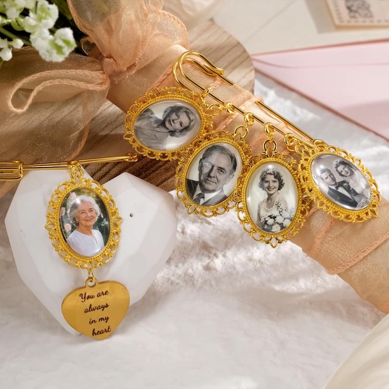May include: Gold-toned memorial pins with oval frames holding black and white photos. One pin has a heart pendant with the inscription "You are always in my heart." The pins are attached to a decorative arrangement with flowers and ribbons.