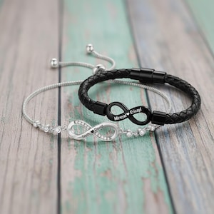 Könnte beinhalten: Zwei Armbänder mit Unendlichkeitszeichen. Eines ist silberfarben mit klaren Steinen und den eingravierten Namen "Smith" und "David". Das andere ist aus schwarzem Leder mit den eingravierten Namen "Mireyal" und "Ericayl". Beide haben verstellbare Verschlüsse.