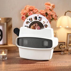 May include: A white retro-style viewer with a black viewing screen and a rotating photo reel. The reel displays several small photos of people. The viewer sits on a wooden surface, with a lit candle and a bouquet of pink flowers in the background.