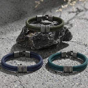 May include: Three rope bracelets in green, navy blue, and teal. Each bracelet has a metal clasp and two small, engraved metal bars. The bracelets are displayed on a textured grey surface.