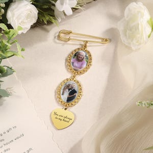 May include: A gold-colored memorial brooch with two oval photo frames and a heart-shaped pendant. The frames hold small photos, and the heart pendant has the inscription "You are always in my heart". The brooch is attached to a safety pin.