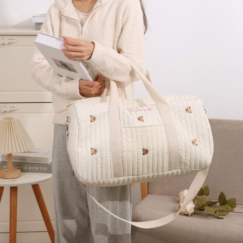 K&ouml;nnte beinhalten: Cremefarbene, gesteppte Reisetasche mit B&auml;renstickerei. Die Tasche hat eine rechteckige Form mit einem oberen Griff und einem langen Schultergurt. Die Tasche hat eine Vordertasche und ist mit kleinen B&auml;rengesichtern verziert.