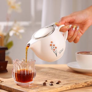 May include: A white ceramic teapot with a silver lid pours a dark liquid into a clear glass. The teapot features a floral design and a handle. A cup of coffee sits on a white saucer. The scene is set on a wooden surface.