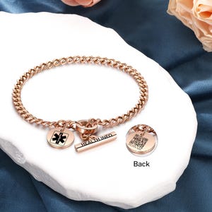 Puede incluir: Pulsera de alerta médica de oro rosa con un dije que presenta un símbolo médico y las palabras "HEALTH INFO" en un lado y un código QR con las palabras "SCAN ME" en el otro.