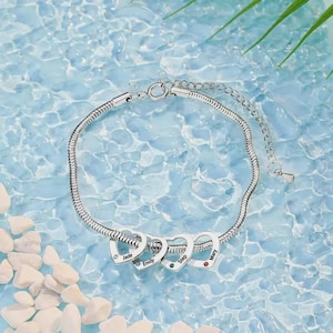 May include: A silver bracelet with a snake chain design. The bracelet features four heart-shaped charms, each engraved with a name: Jade, Emily, Step, and Mary. Each heart has a small gemstone. The bracelet is displayed on a blue water background.