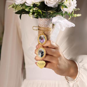 May include: A bridal bouquet with white roses and greenery. A gold charm with two oval photo frames and a heart pendant that reads "You are always in my heart" is attached to the bouquet with a safety pin.