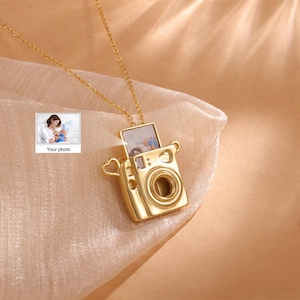 Könnte beinhalten: Goldene Kamera-Anhänger-Kette mit Fotoeinsatz. Der Kamera-Anhänger hat auf jeder Seite ein herzförmiges Detail. Die Kette ist an einer goldenen Kette.