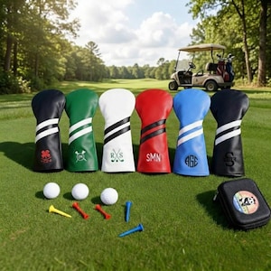 May include: A collection of golf club head covers in various colors, including black, green, white, red, blue, and navy. Each cover features a unique design with stripes and initials. Golf balls, tees, and a small case are also visible on the green grass.