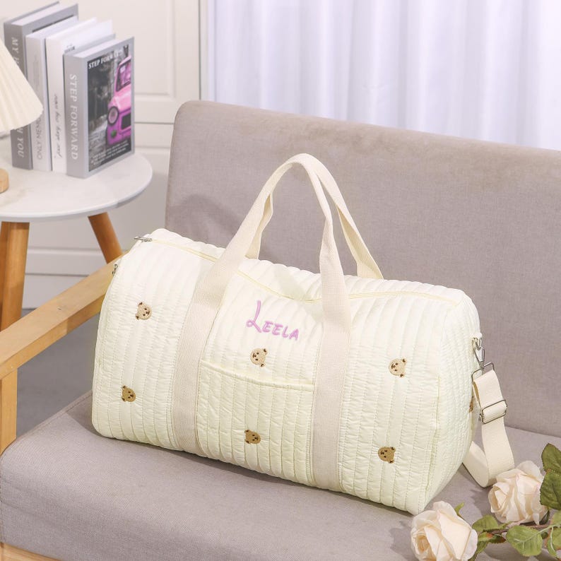 K&ouml;nnte beinhalten: Cremefarbene, gesteppte Reisetasche mit gestickten B&auml;renmotiven und dem Namen "Leela" in Lavendelschrift. Sie hat einen oberen Griff, einen Schultergurt und eine Vordertasche. Die Tasche liegt auf einem beigen Sofa.