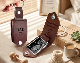 May include: A dark brown leather keyring with a silver metal ring and snap buttons. The exterior is engraved with the word DAD and two hearts. The interior reveals a black and white ultrasound image and the text I need to say I love you Dad.