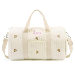 K&ouml;nnte beinhalten: Cremefarbene, gesteppte Reisetasche mit gestickten Teddyb&auml;r-Akzenten. Mit einem oberen Griff, einem abnehmbaren Schultergurt und einer Vordertasche. Der Name "Leela" ist in rosa Schrift auf der Seite gestickt.