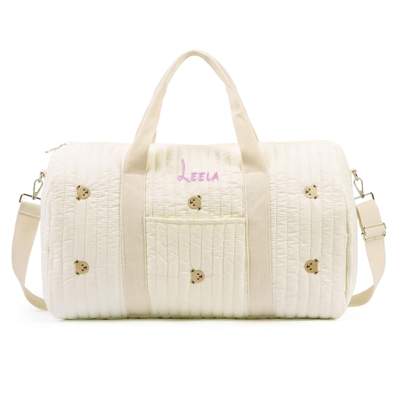 K&ouml;nnte beinhalten: Cremefarbene, gesteppte Reisetasche mit gestickten Teddyb&auml;r-Akzenten. Mit einem oberen Griff, einem abnehmbaren Schultergurt und einer Vordertasche. Der Name "Leela" ist in rosa Schrift auf der Seite gestickt.